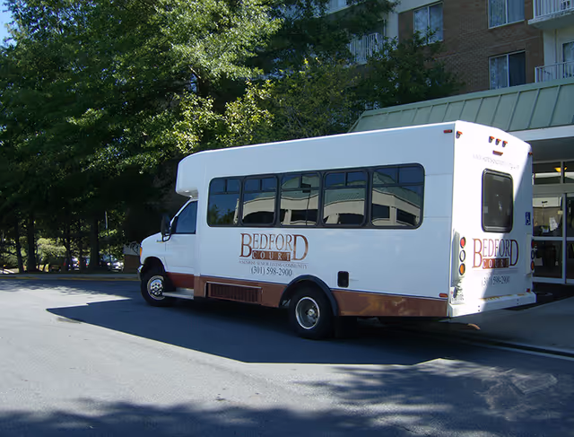 A white Bedford Court shuttle bus parked outside a building entrance with trees and shadows visible in the background.