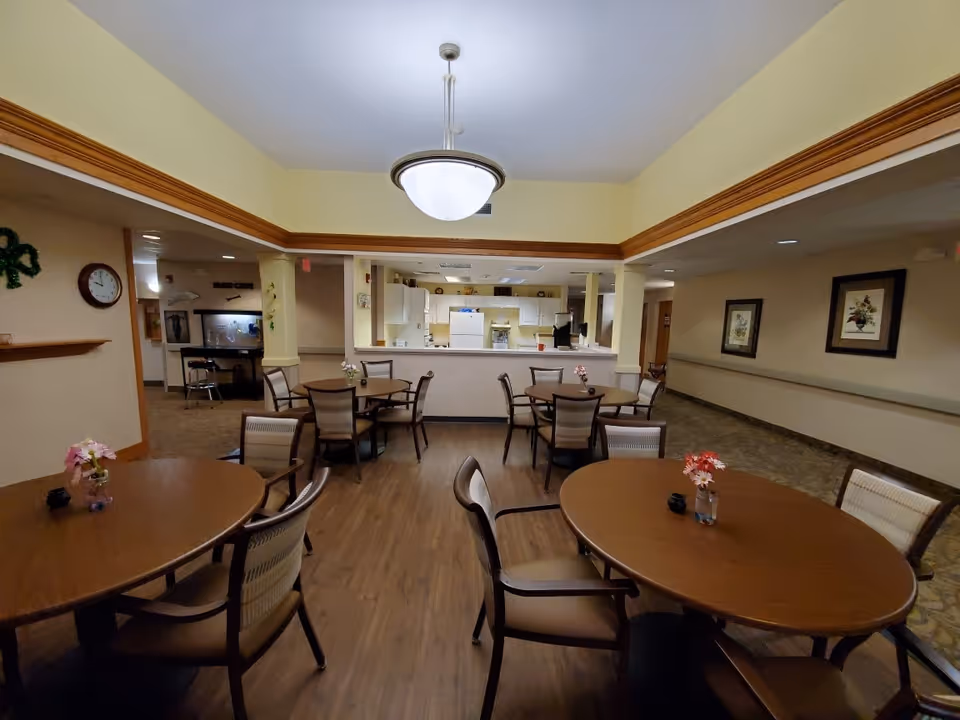 Interior view of a dining area in a senior living facility with several round wooden tables and chairs arranged neatly. Each table has a small flower vase as decoration. The room has wood flooring and beige walls with wooden trim. In the background, there is a kitchen area with white cabinets and appliances visible through a large pass-through window. The ceiling has a central light fixture.