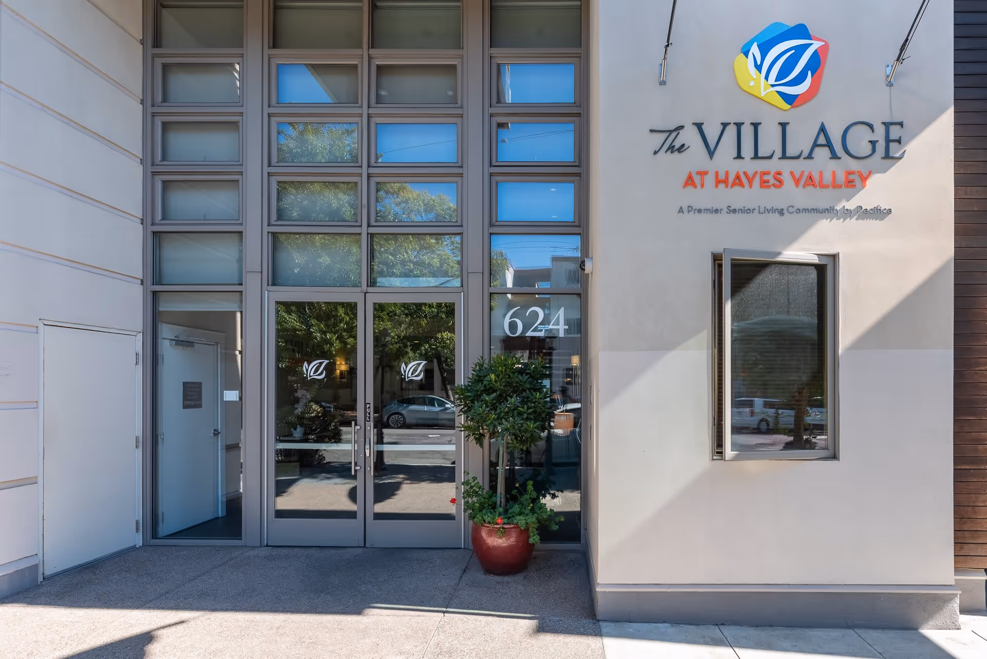 Entrance to The Village at Hayes Valley senior living community with glass double doors, a potted plant, and the facility's name and logo displayed on the exterior wall.