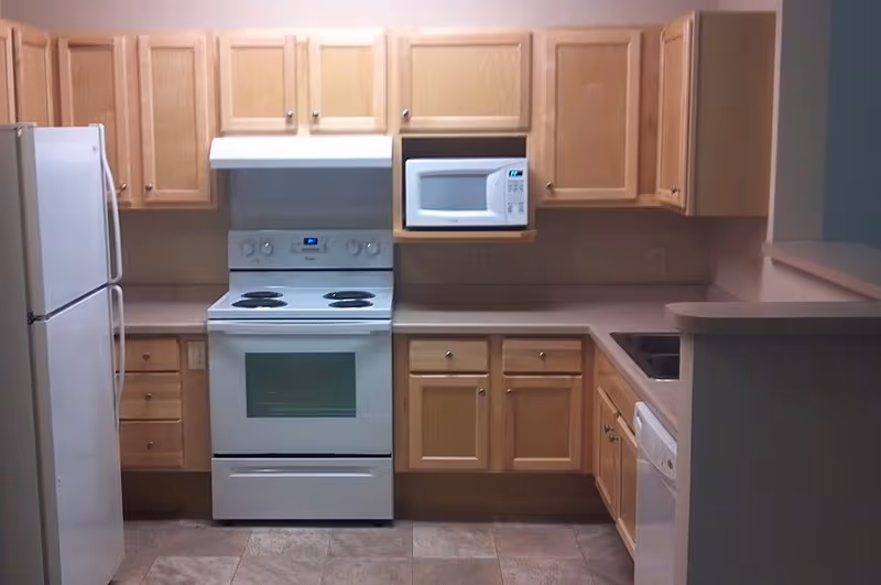 Small L-shaped kitchen with light wood cabinets and white appliances including a refrigerator, stove, microwave, and dishwasher.