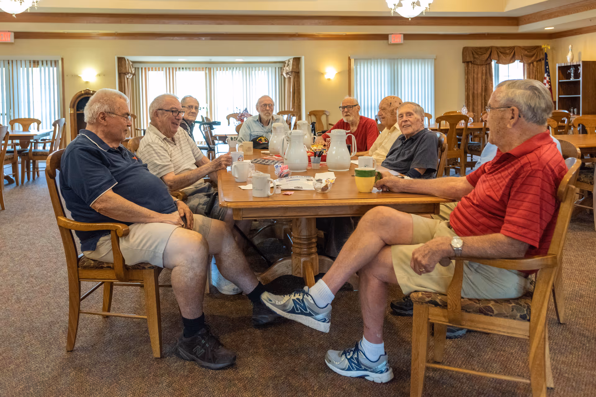 A group of elderly men sitting around a wooden table in a well-lit common room, engaging in conversation and enjoying drinks. The room has large windows with vertical blinds, wooden chairs, and carpeted flooring.