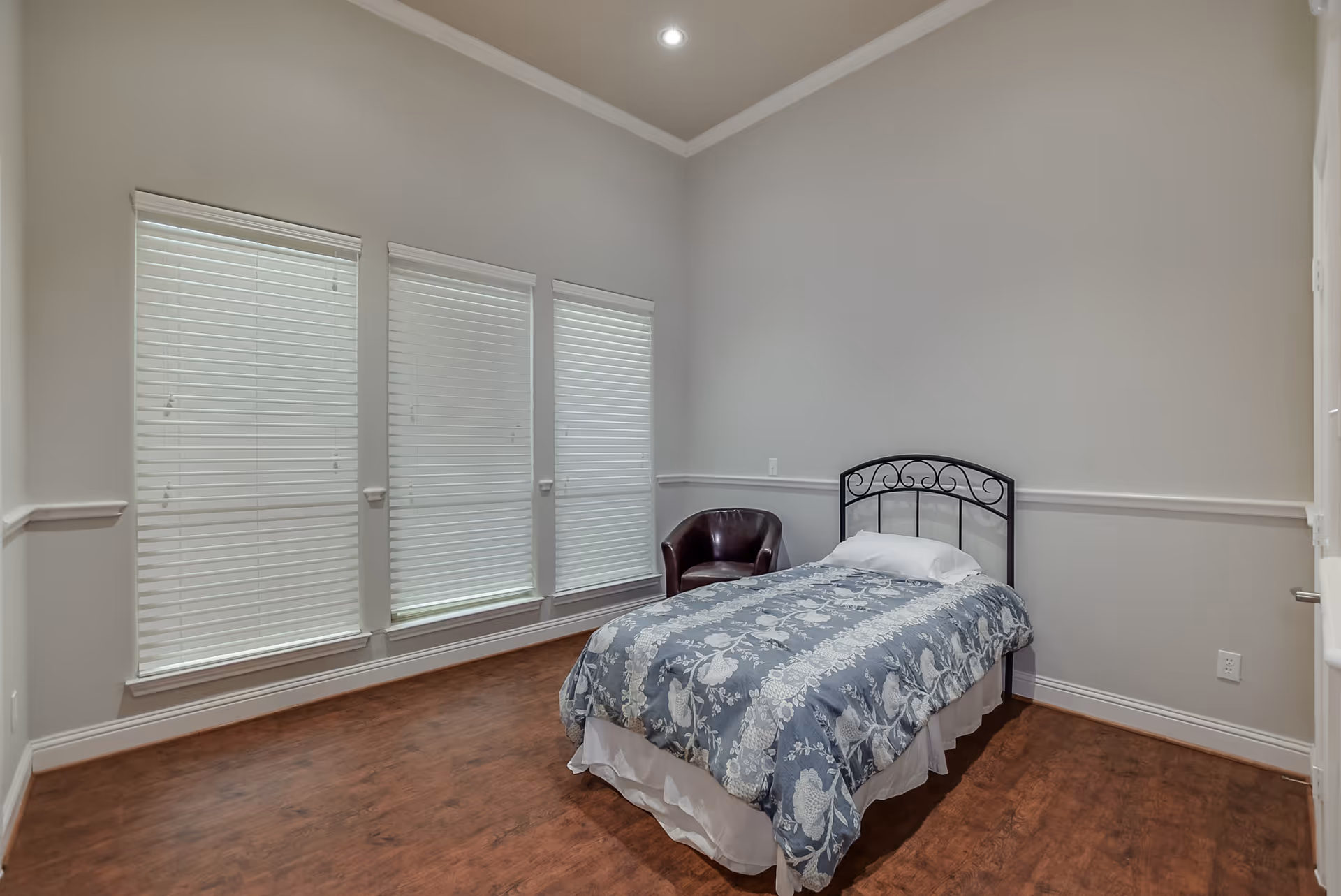 A simple bedroom with a single bed covered with a blue and white floral bedspread, a white pillow, a dark brown leather chair in the corner, three tall windows with white blinds, light gray walls, and wooden flooring.