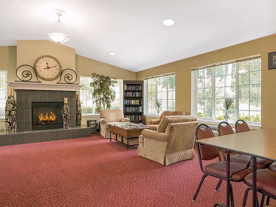A cozy living room area with a red carpet, two beige armchairs, a wooden coffee table with magazines, a fireplace with decorative vases and a large clock above it, a bookshelf filled with books, large windows letting in natural light, and a table with chairs on the right side.