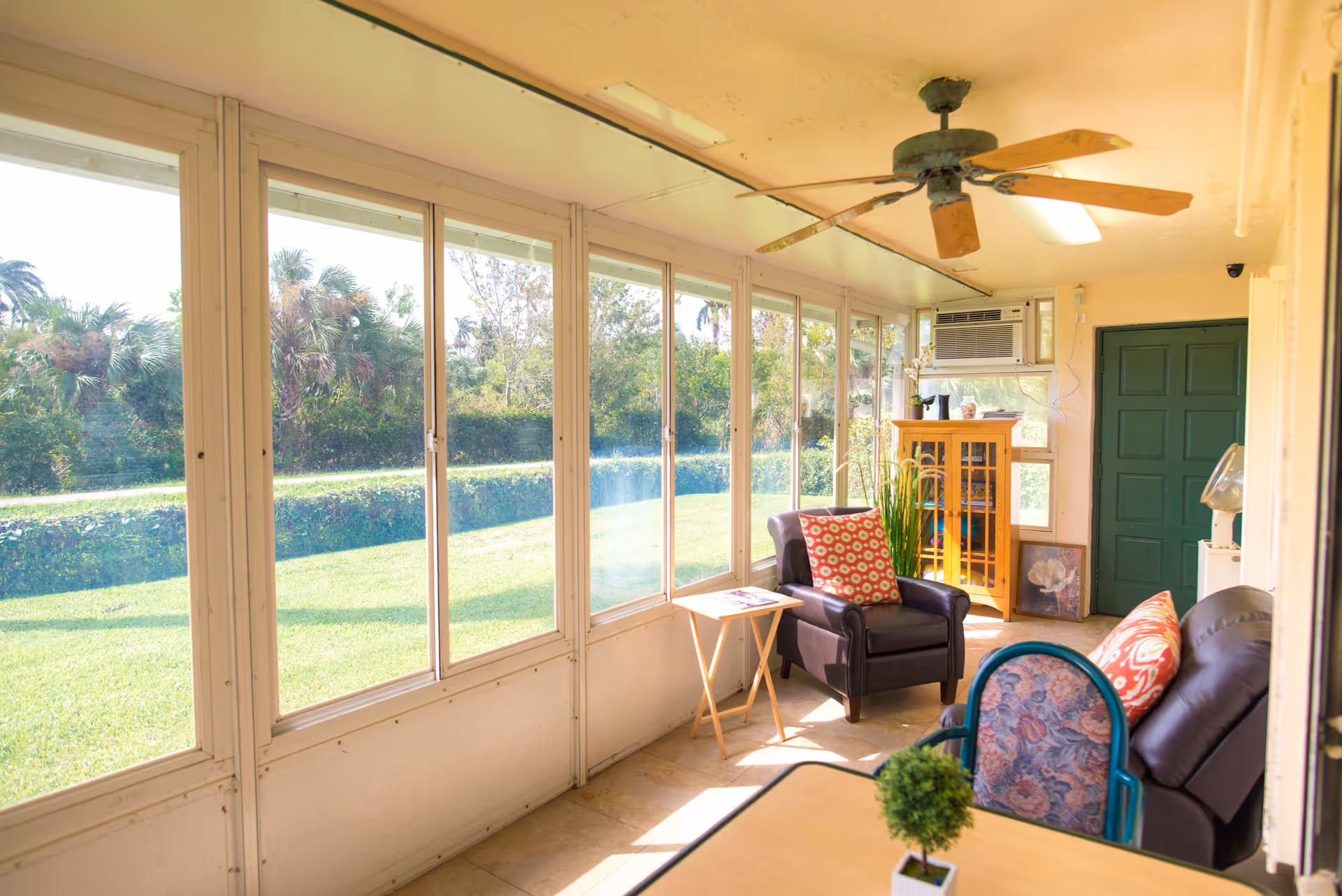 A bright sunroom with large windows overlooking a green lawn and trees. The room contains a ceiling fan, a black armchair with a red patterned pillow, a small wooden side table, a wooden cabinet with glass doors, and a green door. There is also a black leather sofa with a red patterned pillow and a small table with a potted plant in the foreground.