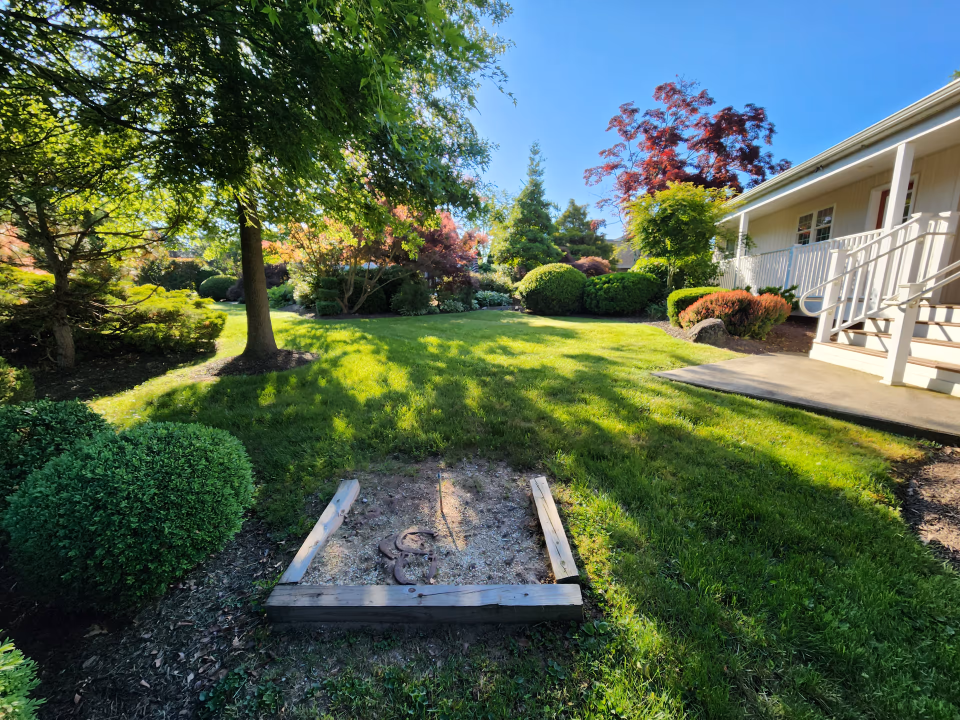 A sunny outdoor garden area with green grass, various bushes, and trees. There is a small wooden horseshoe pit in the foreground and a building with a porch and stairs on the right side.