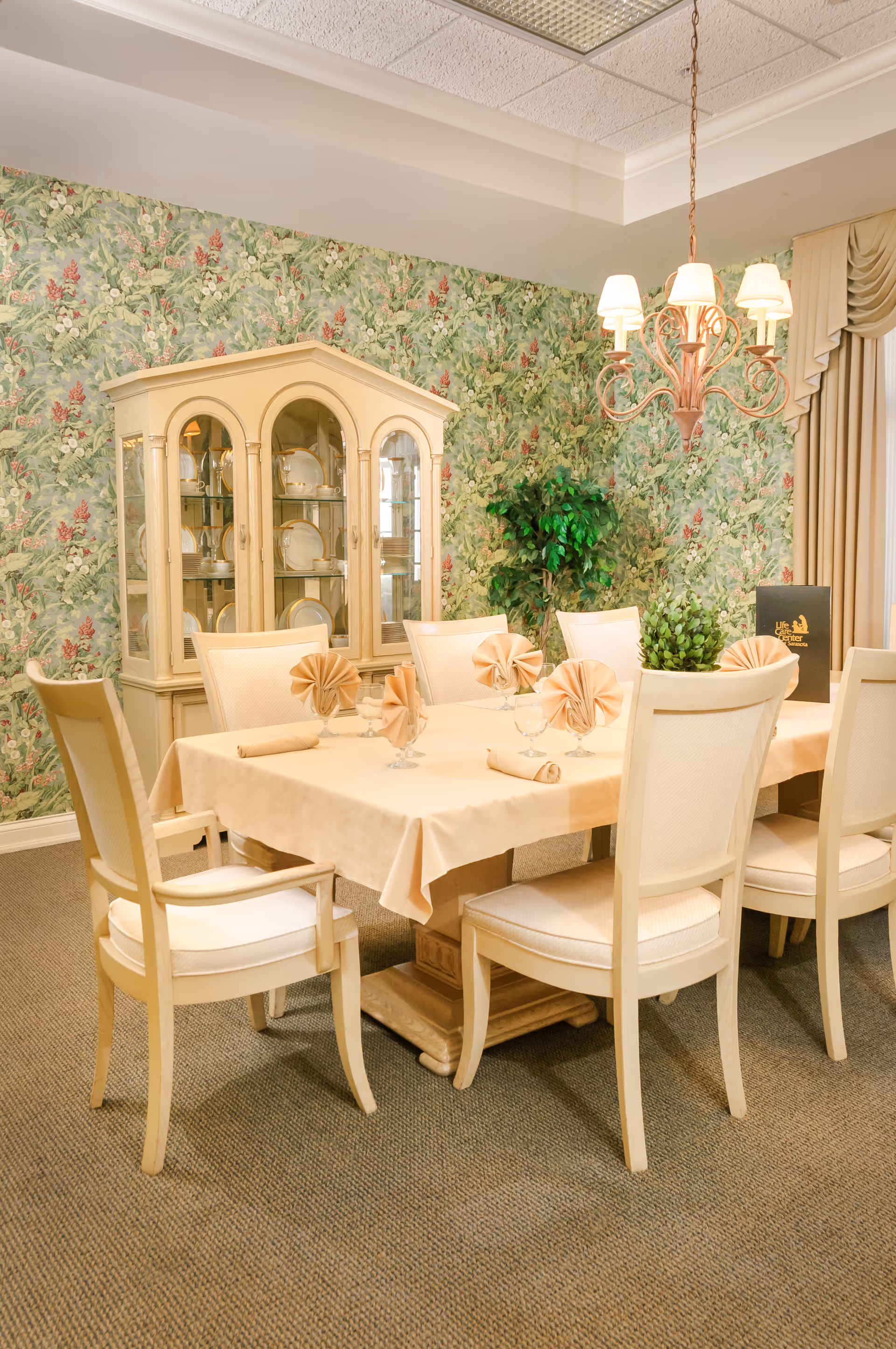A formal dining room with a rectangular table covered with a beige tablecloth, set with folded napkins and glassware. Six cream-colored chairs surround the table. A glass-front china cabinet displaying plates is against a floral wallpapered wall. A chandelier with five lights hangs above the table, and there is a green potted plant in the corner.