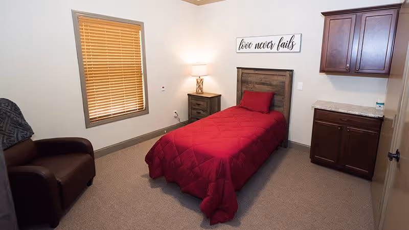 A small bedroom with a single bed covered in a red quilt and a matching pillow. Next to the bed is a wooden nightstand with a lamp. Above the bed is a wall decoration with the words 'love never fails'. There is a window with closed wooden blinds on the left wall, a brown armchair in the corner, and wooden cabinets with a countertop on the right side of the room.