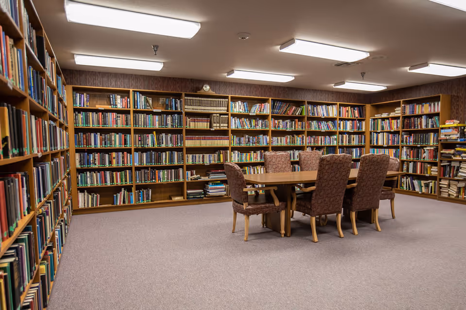A quiet library room with wooden bookshelves filled with books lining the walls. In the center, there is a wooden table surrounded by six upholstered chairs with patterned fabric. The room is carpeted and lit by rectangular fluorescent ceiling lights.