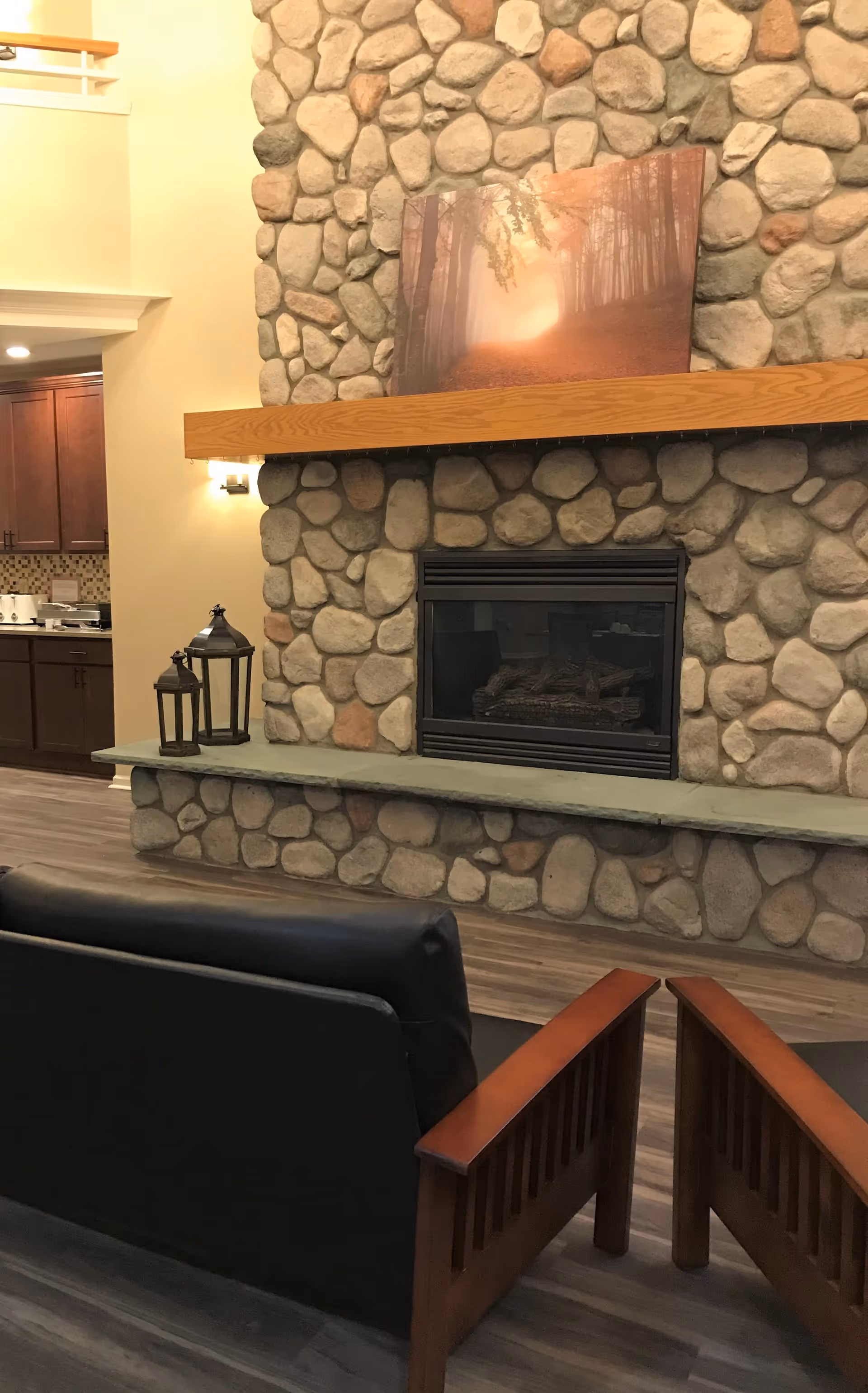 Cozy common room showing a stone fireplace with a wooden mantel, framed artwork, lanterns, and two wooden-framed chairs.