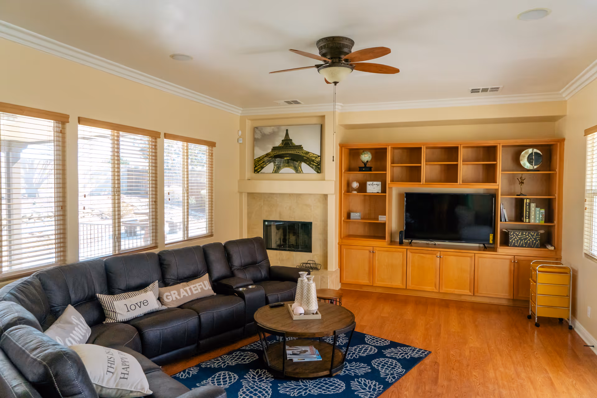 A cozy living room with a large black leather sectional sofa adorned with decorative pillows. There is a round wooden coffee table on a blue rug with a pineapple pattern. The room features a built-in wooden entertainment center with a flat-screen TV, shelves with decorative items, and a fireplace with a framed picture above it. Multiple windows with wooden blinds allow natural light to fill the room. A ceiling fan with wooden blades is mounted on the ceiling.