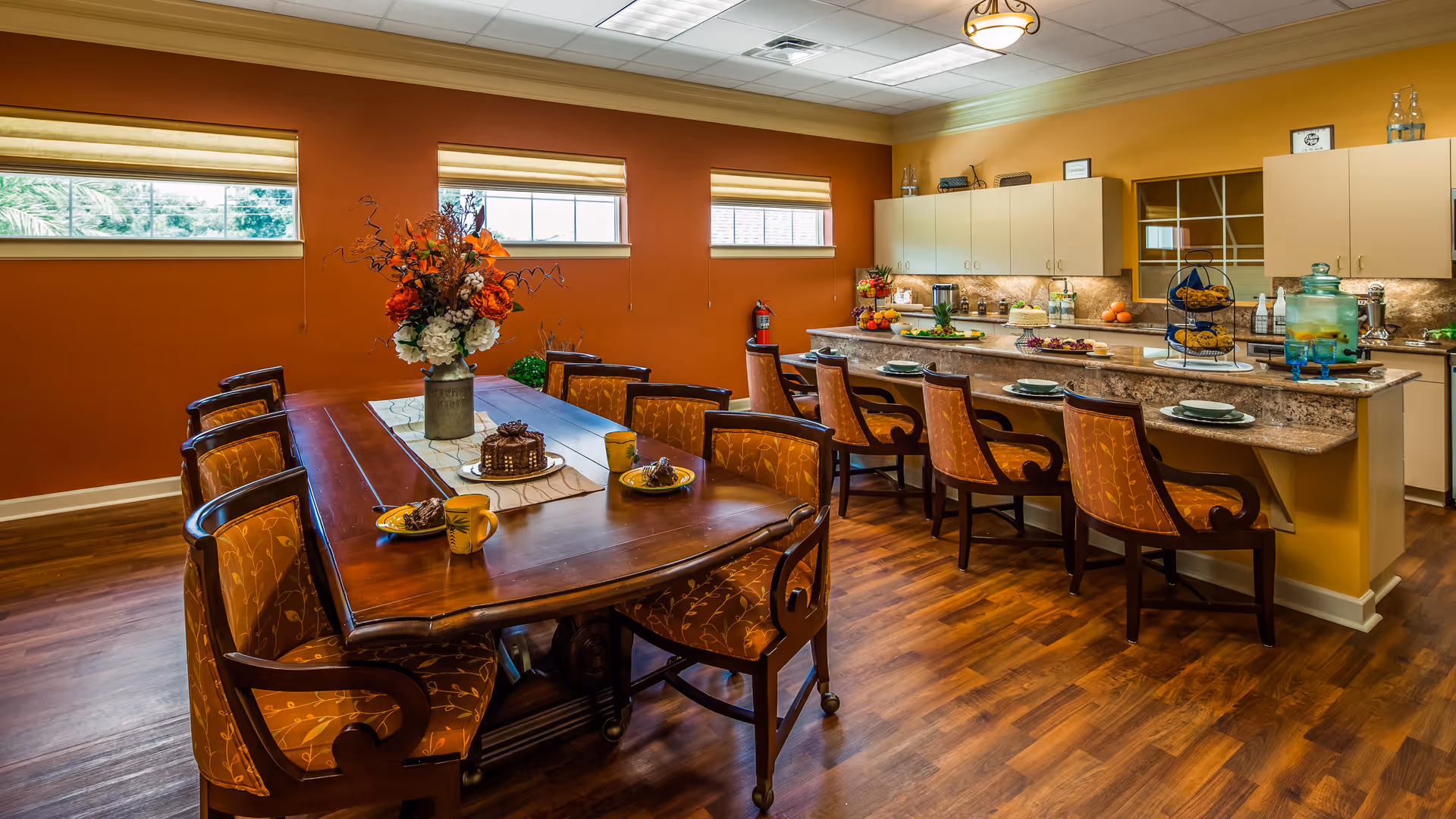 A warm and inviting dining area with a long wooden table surrounded by upholstered chairs. The table is set with plates, mugs, and a centerpiece of flowers. Adjacent to the table is a kitchen counter with additional seating, decorated with plates of food and a large beverage dispenser. The room features orange walls, wooden flooring, and three windows with blinds letting in natural light.