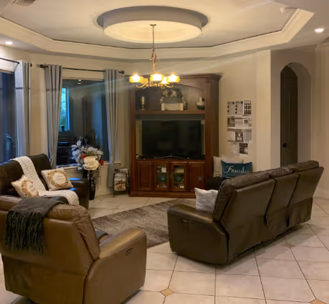Living room with leather recliners arranged around a wooden entertainment center and flat-screen TV under a decorative recessed ceiling and chandelier.
