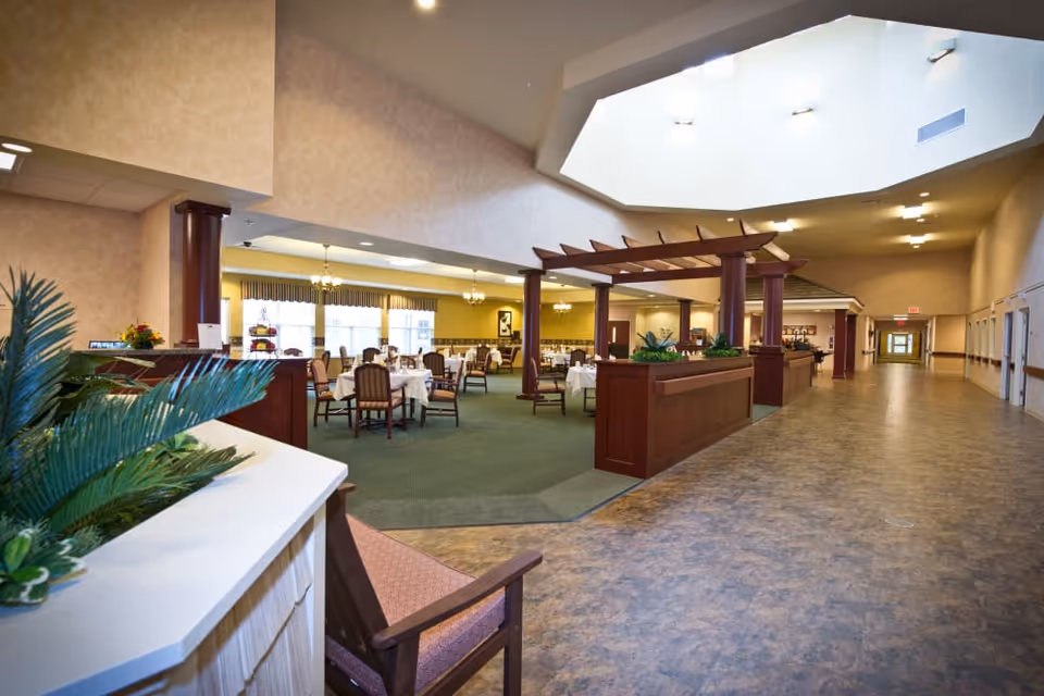 Spacious senior living dining room with set tables and chairs, a wooden pergola-style divider, plants, and a wide corridor under a large skylight.