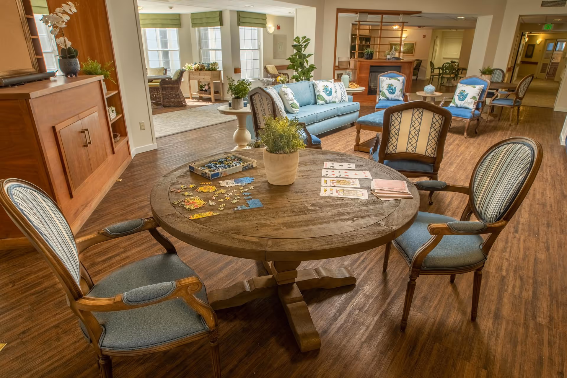 A cozy senior living common area with a round wooden table in the foreground featuring a partially completed jigsaw puzzle, playing cards, and a potted plant. Surrounding the table are four cushioned wooden chairs. In the background, there is a seating area with a blue sofa, armchairs with floral cushions, a fireplace, and large windows letting in natural light.
