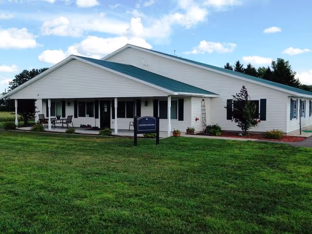 Single-story white building with a green roof, covered front porch, sign, and a large lawn under a blue sky.