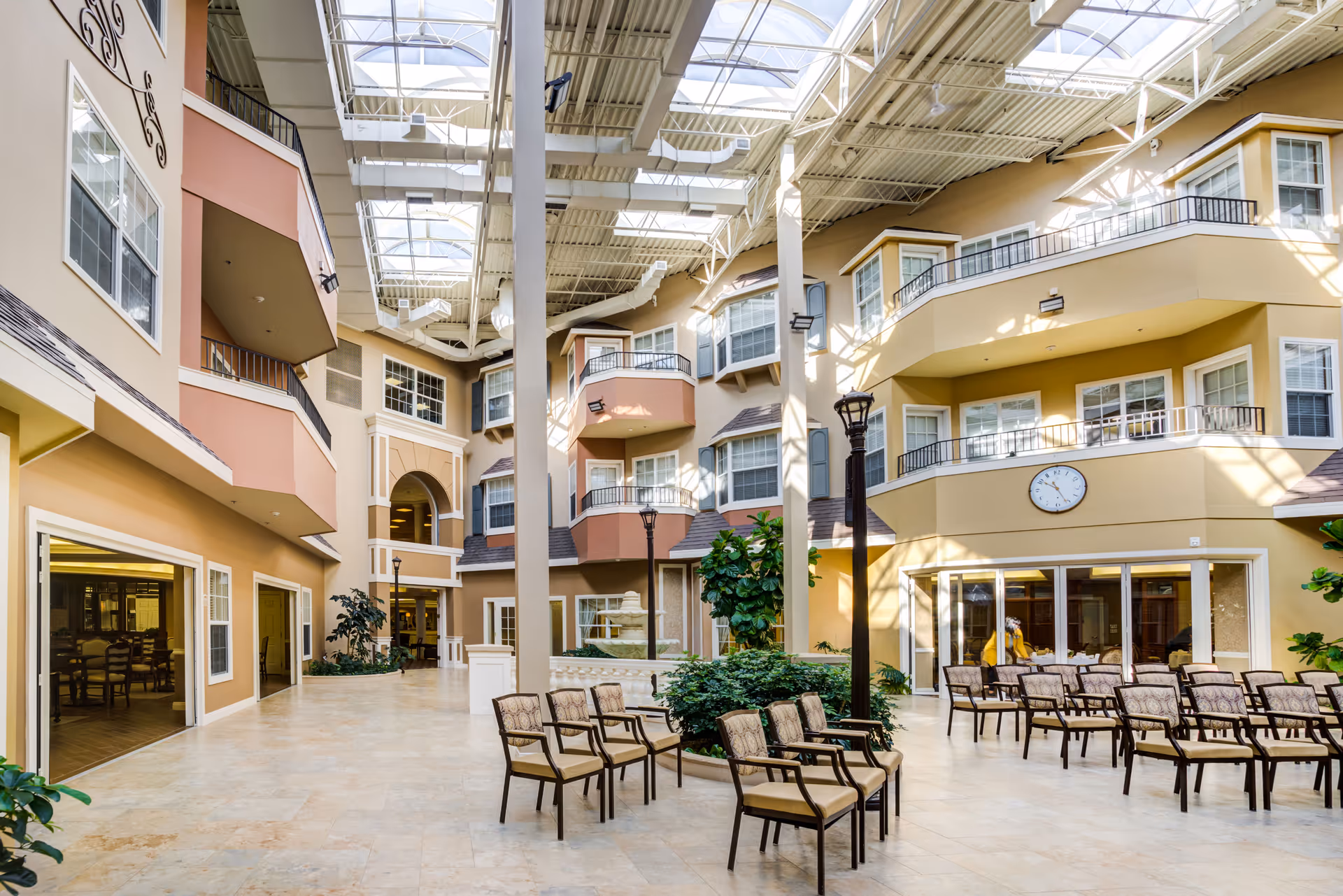 A spacious indoor atrium area with multiple floors of residential units featuring balconies and large windows. The space has a high ceiling with skylights allowing natural light to fill the area. Several chairs are arranged in rows around a central planter with greenery. The walls are painted in warm tones of beige and peach, and there is a clock mounted on one wall. The area appears to be part of a senior living facility.