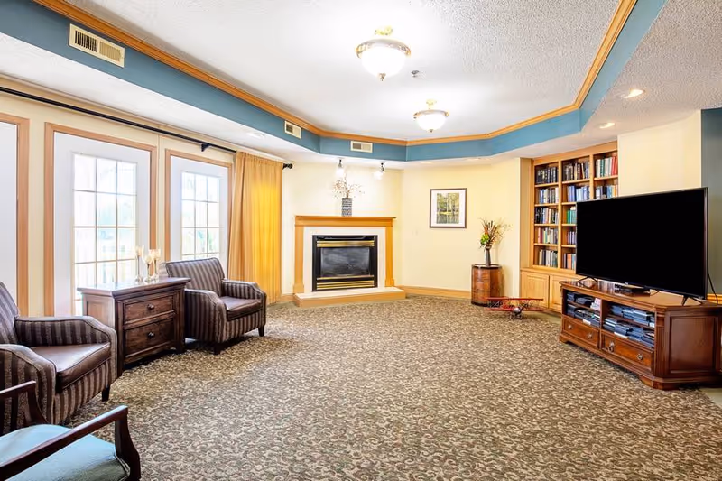 A cozy living room with patterned carpet, two striped armchairs next to a wooden side table, a fireplace with a wooden mantle, a built-in bookshelf filled with books, a large flat-screen TV on a wooden TV stand, and a framed picture on the wall. The room has large windows with yellow curtains and ceiling lights.