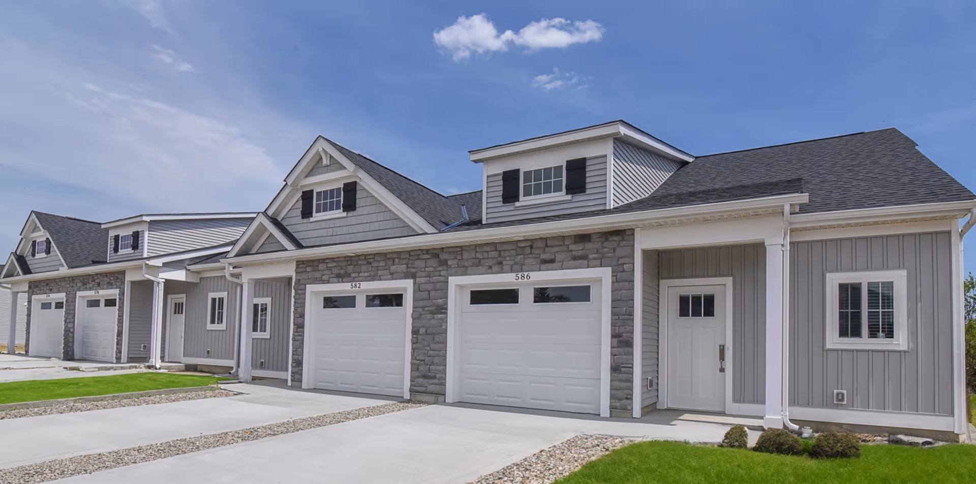 Exterior view of a row of modern townhomes with gray siding, stone accents, white garage doors, and small front lawns under a blue sky with a few clouds.