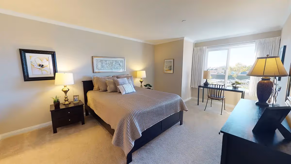 A bright and neatly arranged bedroom with a large bed covered in beige bedding and multiple pillows. There are two bedside tables with lamps on either side of the bed. A framed picture hangs above the bed. A window with white curtains lets in natural light, illuminating a small black desk and chair positioned in front of it. The room has light-colored walls and carpeted flooring.
