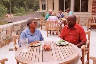 Two elderly people sitting at a round wooden table outdoors, enjoying a meal and conversing. In the background, other elderly individuals are seated at similar tables near a building with large windows, surrounded by greenery.