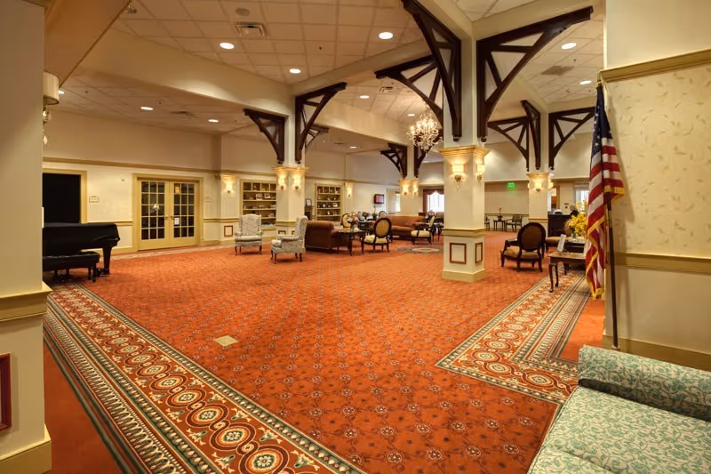 Spacious and elegant common area with red patterned carpet, cream-colored walls, and dark wooden beams on the ceiling. The room is furnished with various seating arrangements including armchairs, sofas, and small tables. There is a grand piano on the left side and an American flag on the right side near a patterned sofa. The area is well-lit with ceiling lights and wall sconces, and there are built-in shelves along the far wall.