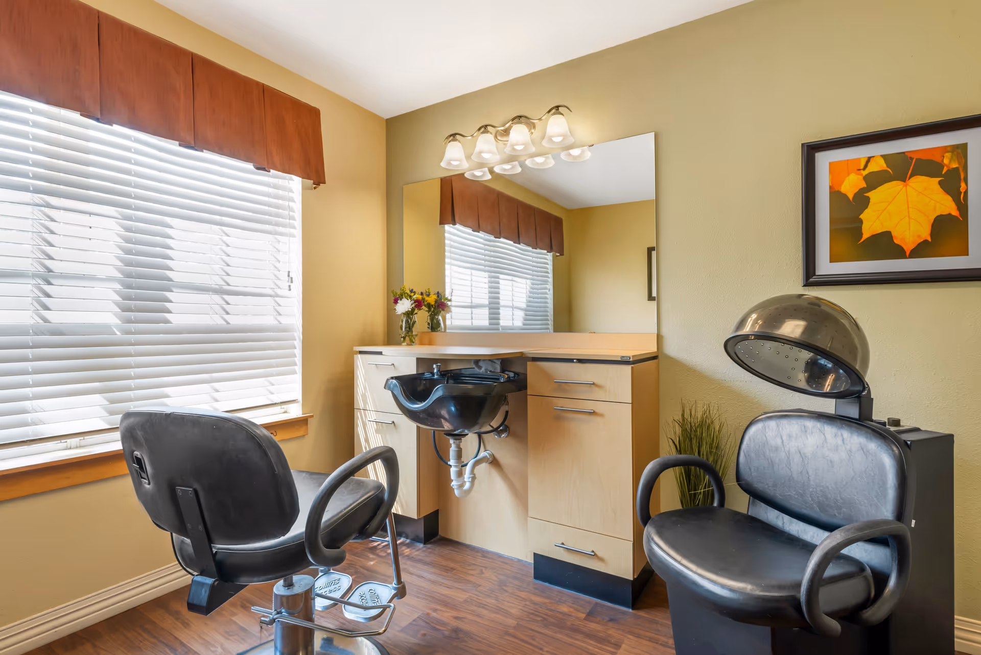 A small hair salon area with two black salon chairs, a black hair washing sink attached to a wooden cabinet with drawers, a large mirror above the sink, a window with white blinds and a brown valance, a wall-mounted light fixture with four bulbs, a framed picture of autumn leaves on the wall, and a small vase with flowers on the cabinet.