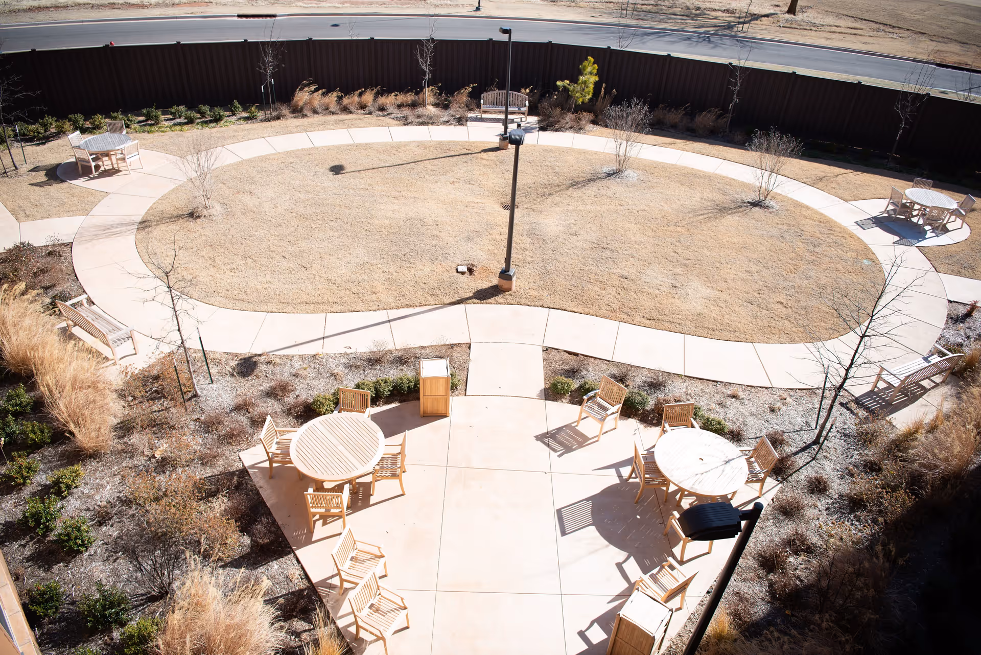 A circular outdoor courtyard with a paved walking path surrounding a dry grass area. Several wooden tables and chairs are arranged on the paved area, along with benches and small trees and shrubs planted around the perimeter. A tall fence and a road are visible in the background.