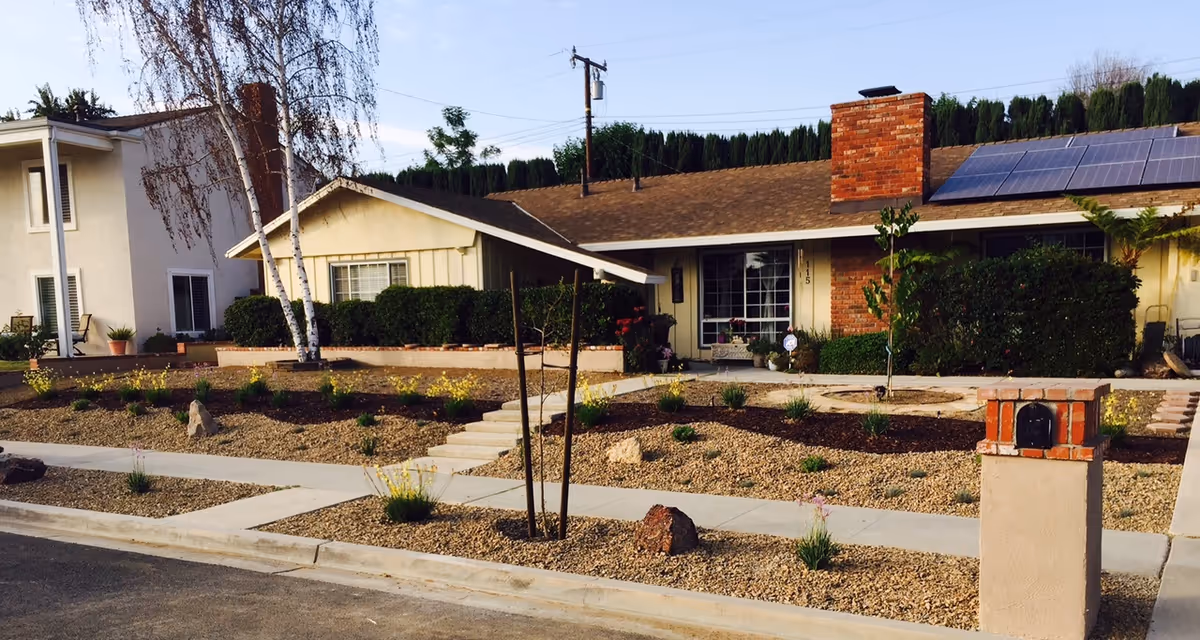 Front exterior view of a single-story residential building with a brick chimney, solar panels on the roof, and a landscaped front yard featuring gravel, small plants, and a concrete walkway leading to the entrance. A mailbox is visible near the street curb.