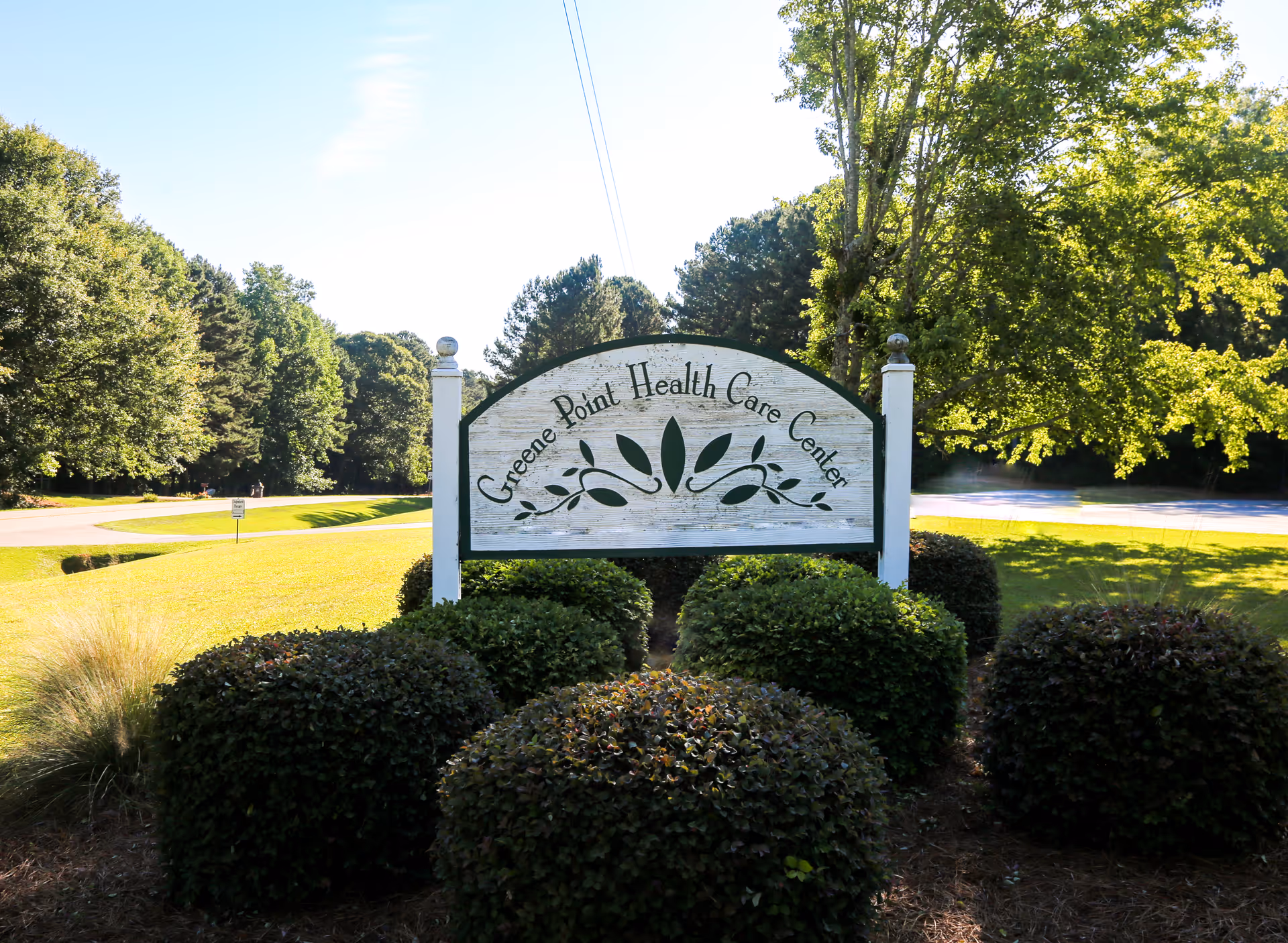 Outdoor view of a white wooden sign with green trim that reads 'Greene Point Health Care Center' surrounded by neatly trimmed bushes and trees in the background under a clear sky.