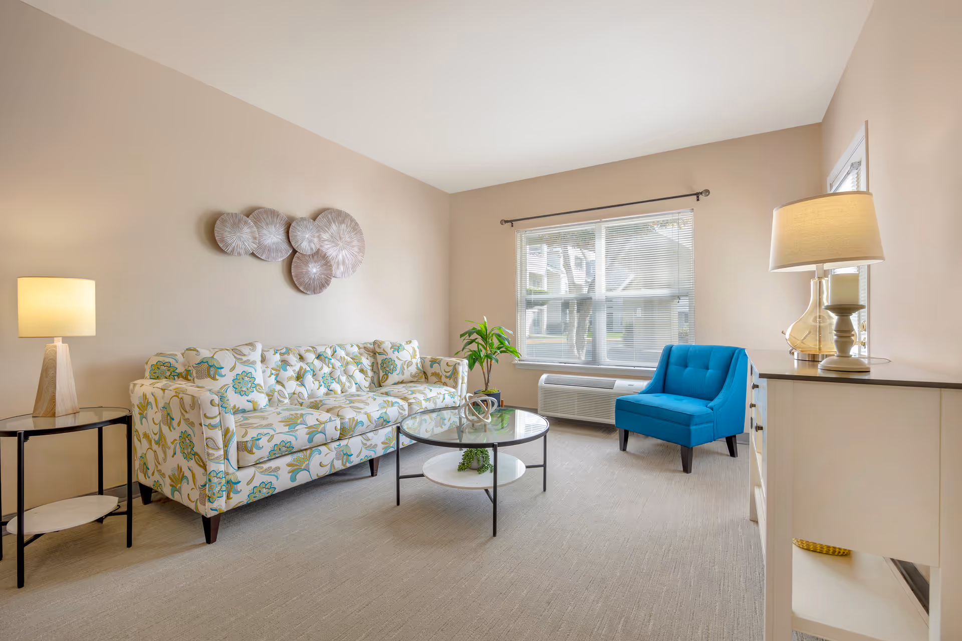 A bright living room with a floral patterned sofa, a blue armchair, a round glass coffee table, a side table with a lamp, a white dresser with a lamp and candle, and a large window with blinds letting in natural light.