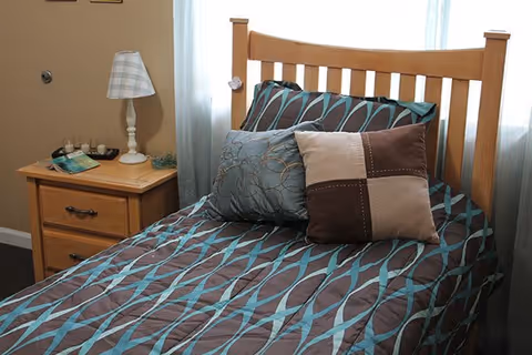 A neatly made single bed with a wooden headboard, featuring a brown and teal patterned bedspread and three decorative pillows. Next to the bed is a wooden nightstand with two drawers, a white lamp with a plaid lampshade, and some small decorative items. A window with sheer white curtains is behind the bed.