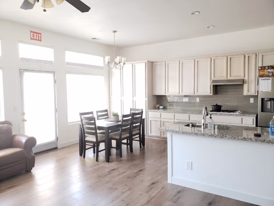 Bright and modern kitchen and dining area with white cabinets, granite countertops, a dining table with six chairs, a ceiling fan, and a leather armchair near a door with large windows letting in natural light.
