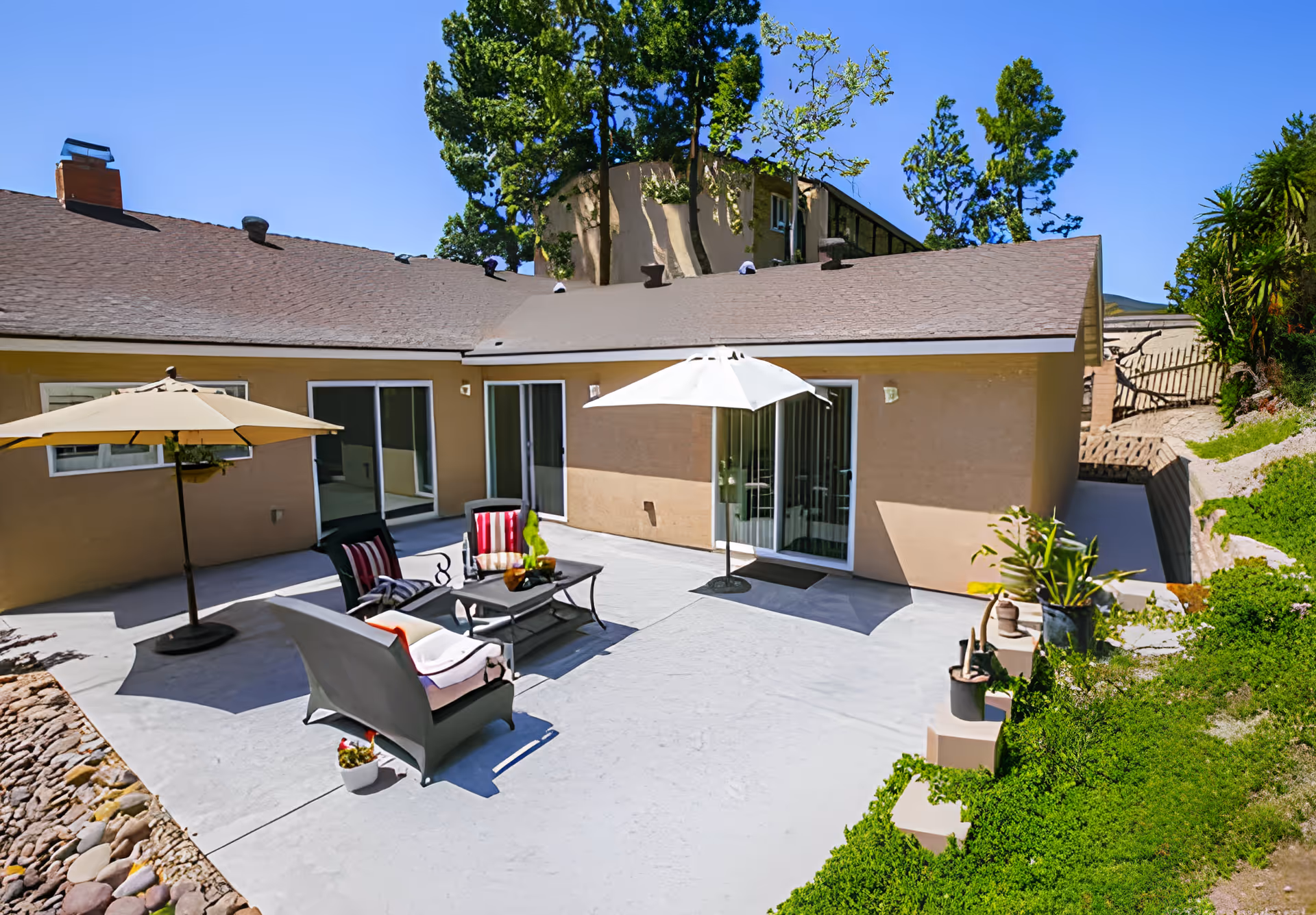Outdoor patio area at Mount Carmel Assisted Living featuring a concrete floor, two umbrellas providing shade, several cushioned chairs with striped pillows, a coffee table with decorative items, and surrounding greenery and plants.