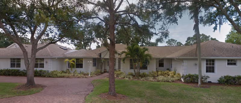 Exterior view of a single-story residential building with a light-colored roof and white walls, surrounded by trees and landscaped greenery, with a brick-paved driveway leading to the entrance.