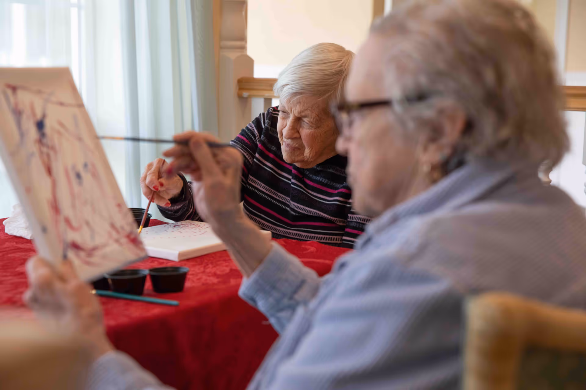 Two elderly women sitting at a table covered with a red tablecloth, engaged in painting on canvases with paintbrushes and small paint containers.