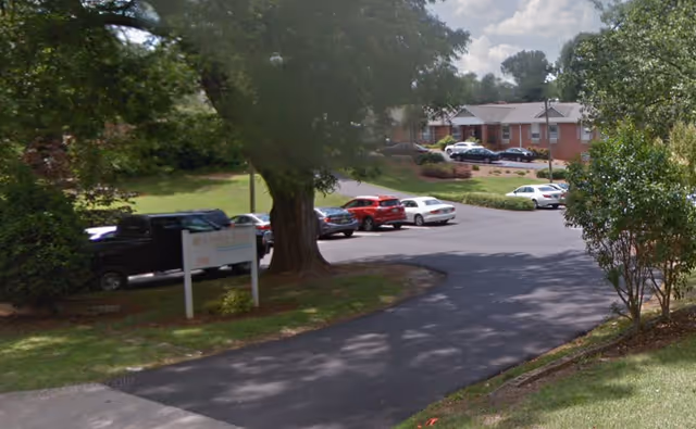 Driveway and parking lot in front of a single-story brick building with parked cars, trees, and a grassy lawn.