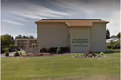 Exterior view of a beige two-story building with a brown roof, labeled Ponderosa Retirement Assisted Living, surrounded by a well-maintained lawn and landscaped garden beds under a clear sky.