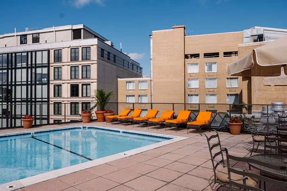 Rooftop swimming pool with orange lounge chairs, potted plants, and surrounding city buildings under a blue sky.