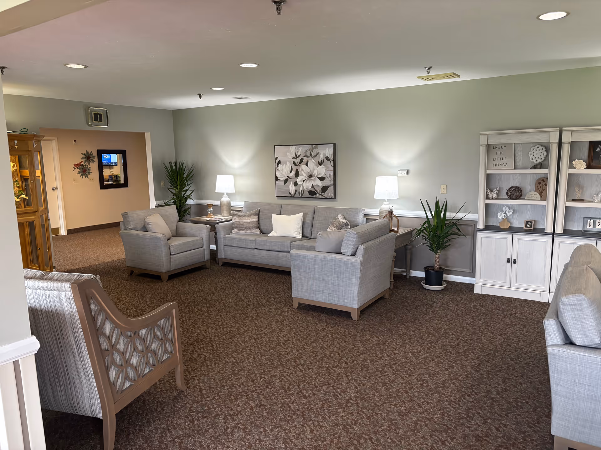 A cozy living room area in an assisted living facility with light gray upholstered sofas and armchairs arranged around a small wooden table with a lamp. The walls are painted light green with white trim, and there is a large floral painting hanging above the sofa. Two potted plants are placed near the walls, and a white wooden cabinet with decorative items and a sign that reads 'ENJOY THE LITTLE THINGS' is visible on the right side. The floor is carpeted in a brown pattern, and recessed ceiling lights illuminate the space.