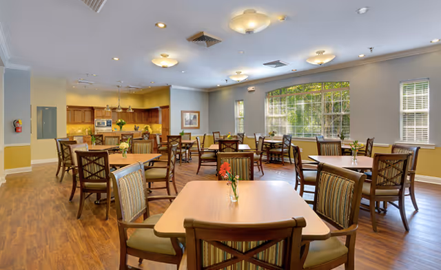 A spacious dining room with multiple wooden tables and chairs arranged neatly. Each table has a small vase with flowers. Large windows on one side let in natural light, and a kitchen area with wooden cabinets and appliances is visible in the background.