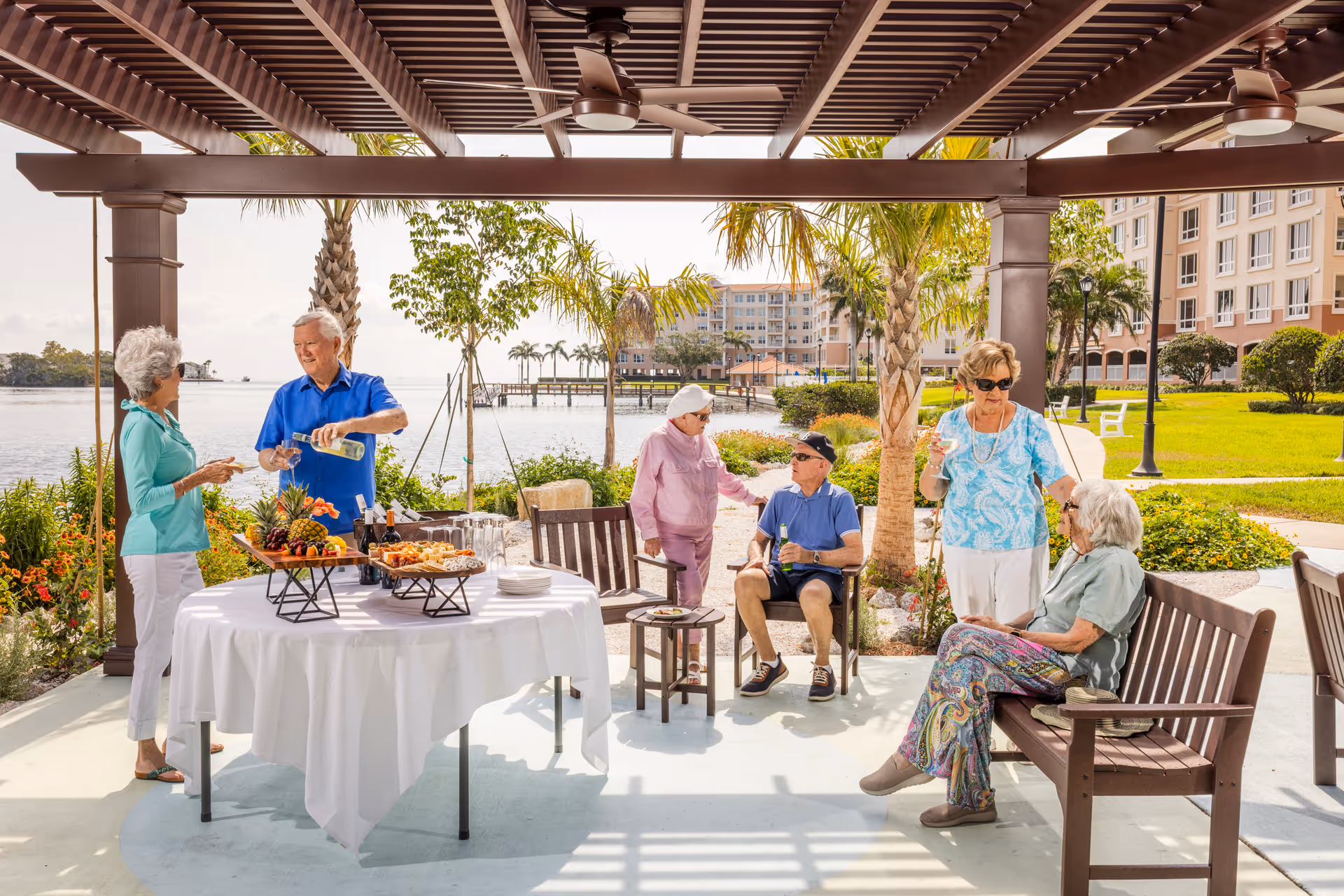 A group of six elderly people socializing outdoors under a wooden pergola near a waterfront. There is a table with a white tablecloth holding food and drinks. Palm trees and a large building are visible in the background on a sunny day.
