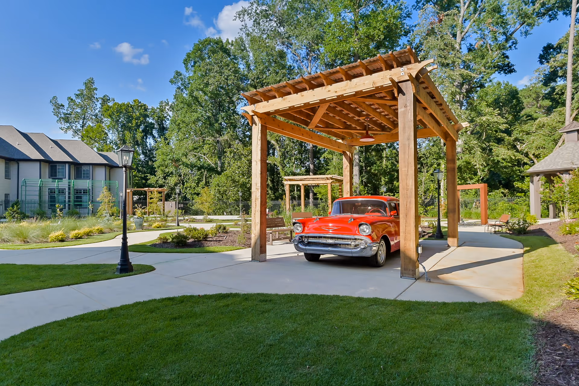A bright red vintage car parked under a wooden pergola in a landscaped outdoor area with green grass, paved walkways, lamp posts, and trees in the background on a sunny day.