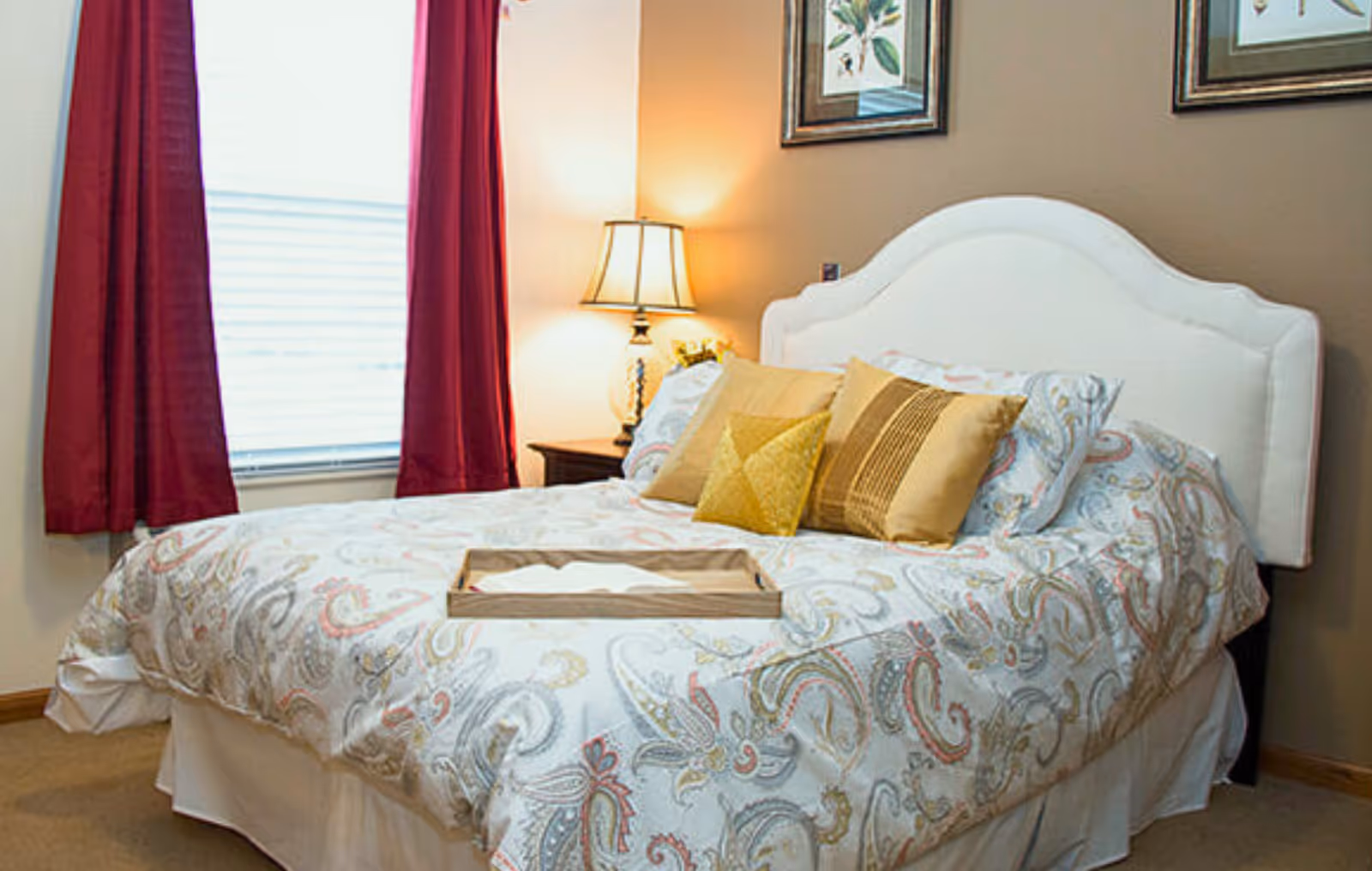 A cozy bedroom with a white upholstered headboard, a bed covered with a paisley-patterned comforter and several decorative pillows in shades of yellow and gold. There is a wooden nightstand with a lamp emitting warm light next to the bed, and a window with red curtains letting in natural light. Two framed botanical prints hang on the wall above the bed.