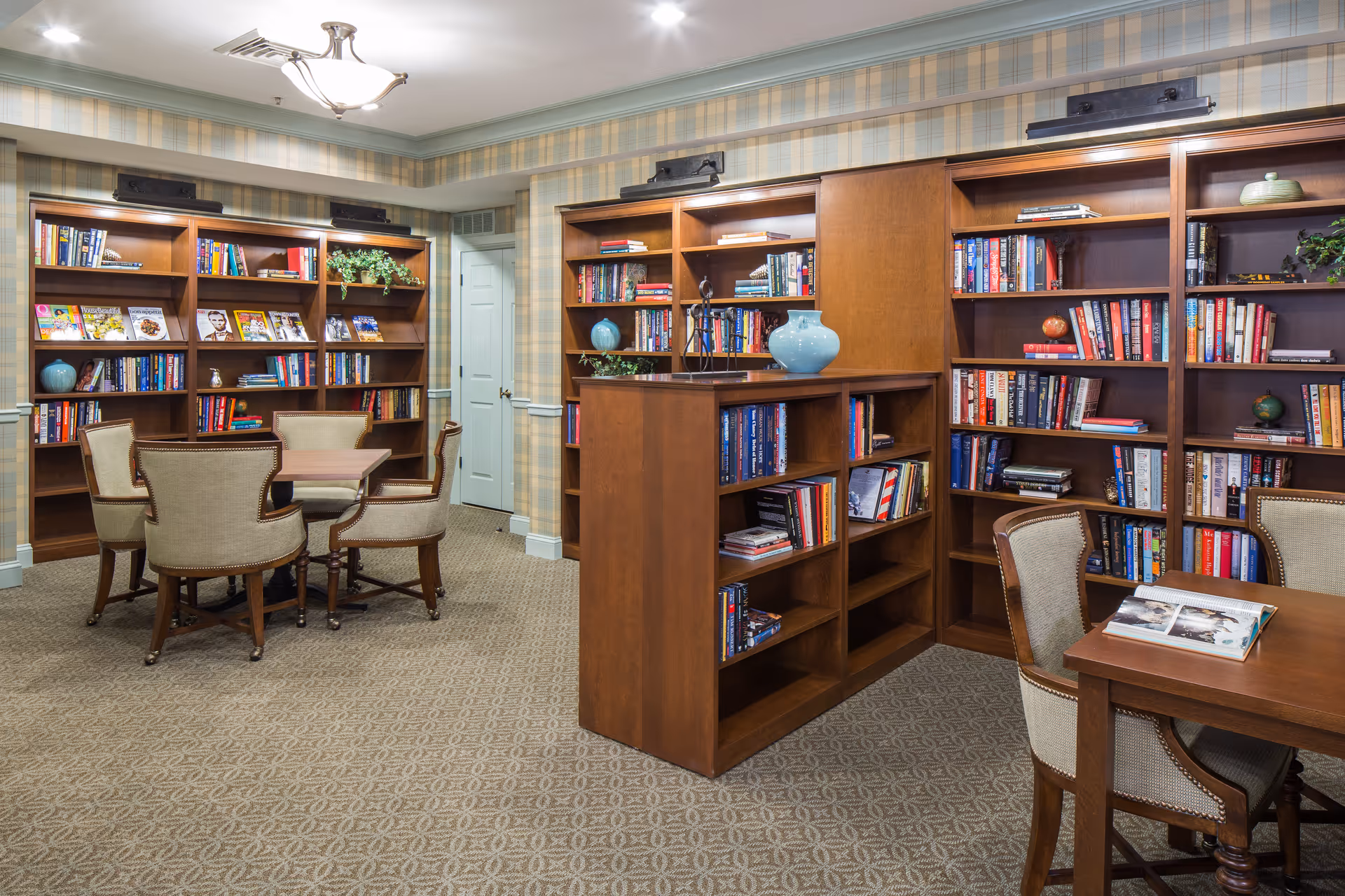A cozy library room with wooden bookshelves filled with books and decorative items. There are two wooden tables with upholstered chairs around them. The walls have a plaid wallpaper and the carpet has a subtle pattern. A ceiling light fixture illuminates the room.