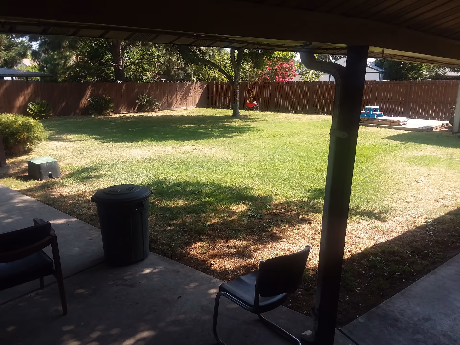 Covered patio looking out onto a grassy fenced backyard with chairs, a trash can, and children's play equipment.