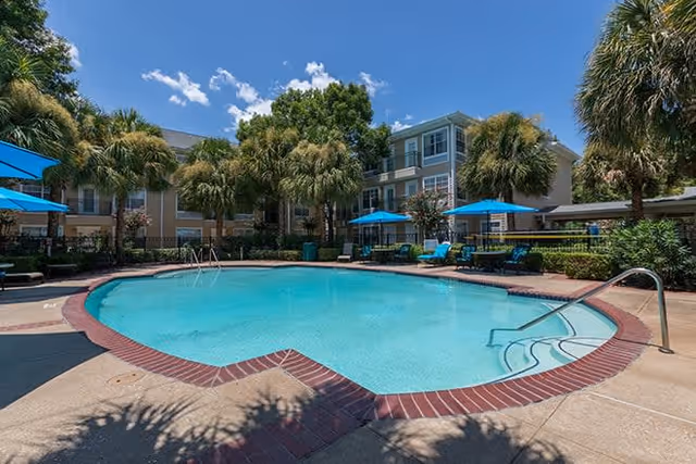 Outdoor swimming pool surrounded by palm trees and lounge chairs with blue umbrellas, adjacent to a multi-story residential building under a clear blue sky.
