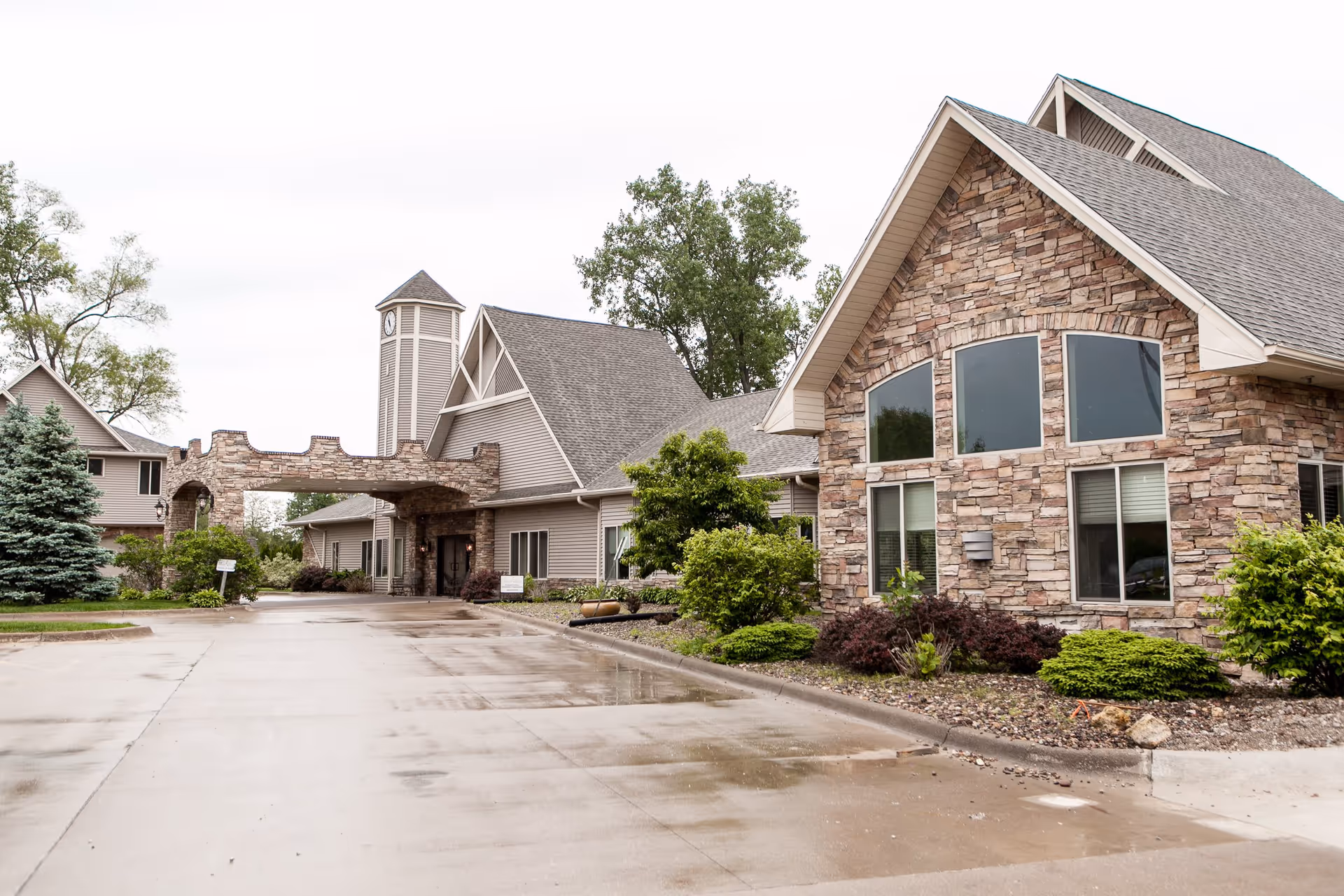 Exterior view of a senior living facility building with stone and siding facade, large windows, a clock tower, and a covered entrance. The driveway is wet, and there are landscaped bushes and trees around the building.