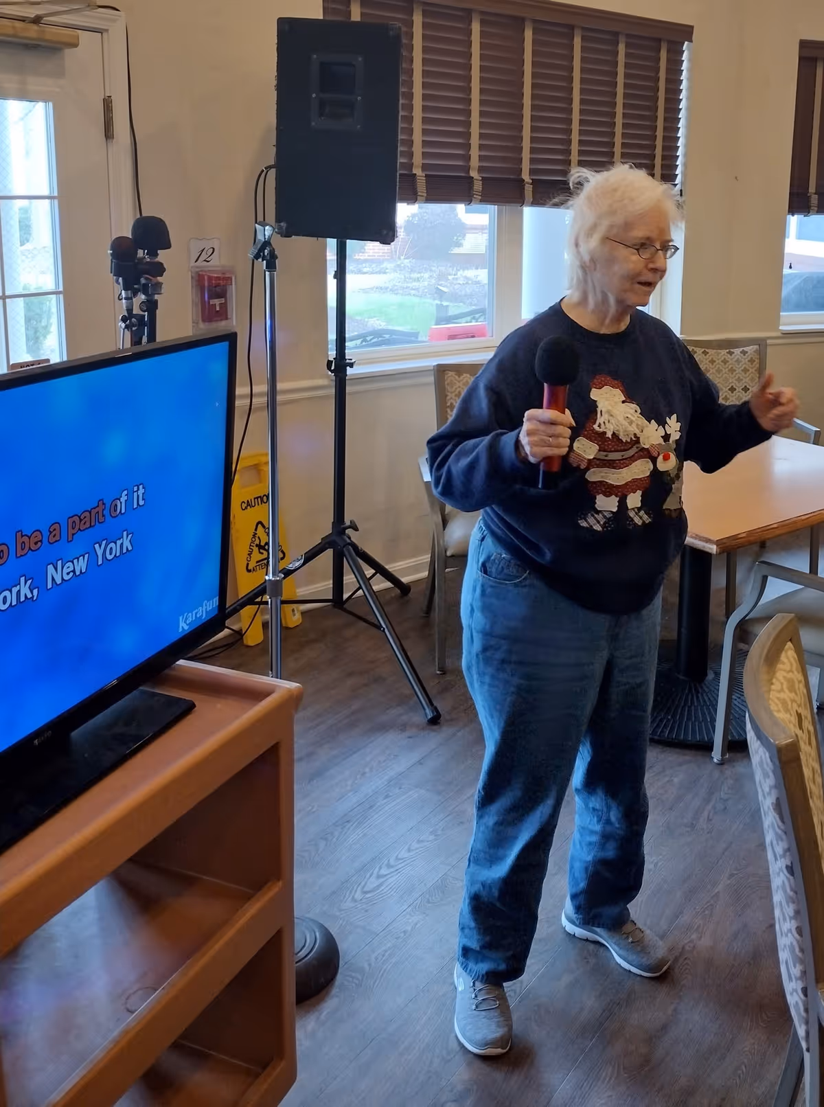 An elderly woman wearing a Christmas-themed sweater and glasses is holding a microphone and appears to be singing or speaking in a room with wooden floors, tables, and chairs. A large TV screen displaying karaoke lyrics and a speaker on a stand are visible nearby. The room has windows with brown blinds and a door with a window.