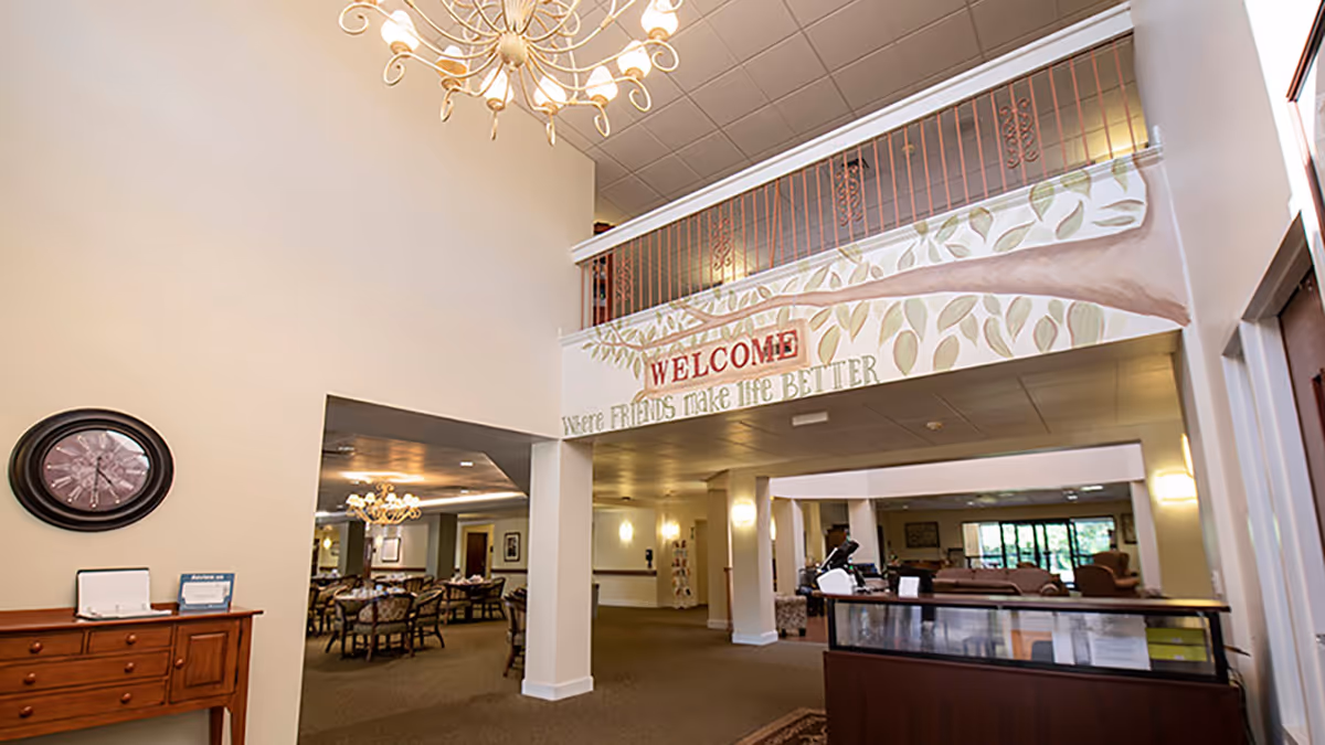 Interior view of a senior living facility lobby with a high ceiling and chandelier. A decorative sign on the upper wall reads 'WELCOME where FRIENDS make life BETTER.' There is a wooden reception desk on the right and a dining area with tables and chairs visible through an open doorway on the left.