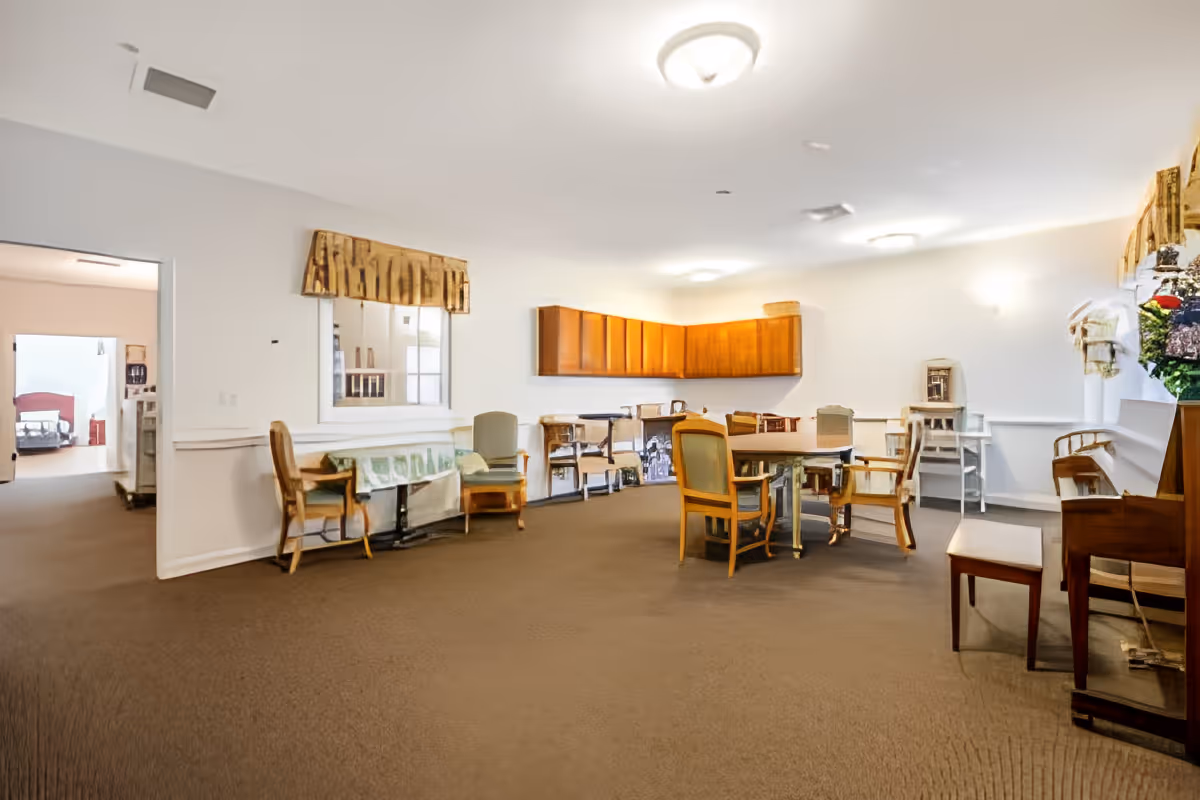 A spacious room with beige carpet and white walls, furnished with wooden chairs and tables arranged in the center. There are wooden cabinets mounted on the far wall, a piano on the right side with a floral arrangement on top, and a window with a valance on the left wall. The room is well-lit with ceiling lights, and an adjacent room with a bed is visible through an open doorway on the left.